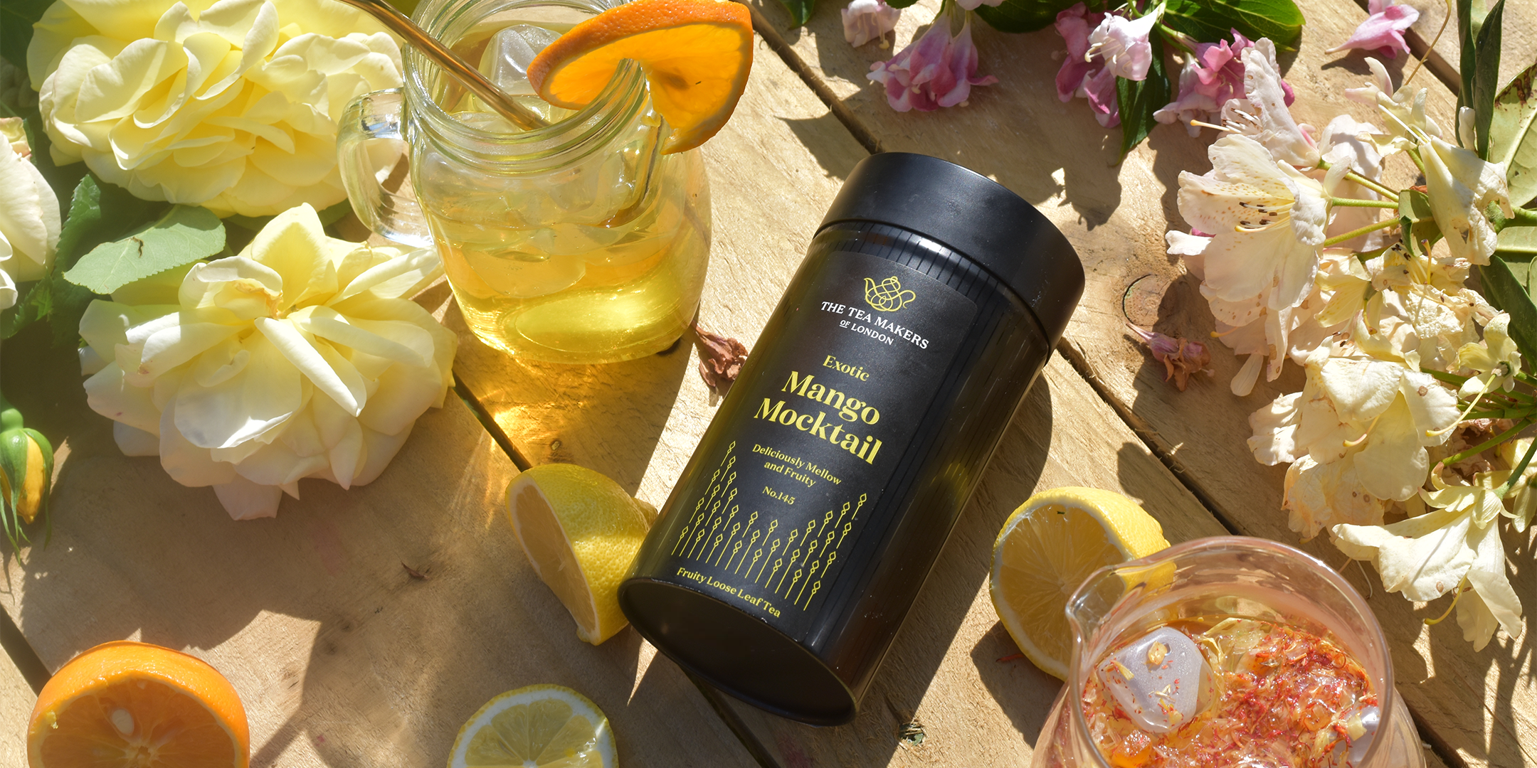 A black tin of fruit tea, surrounded by fruit tea in mugs, slices of lemon and flowers on a wooden picnic table. 